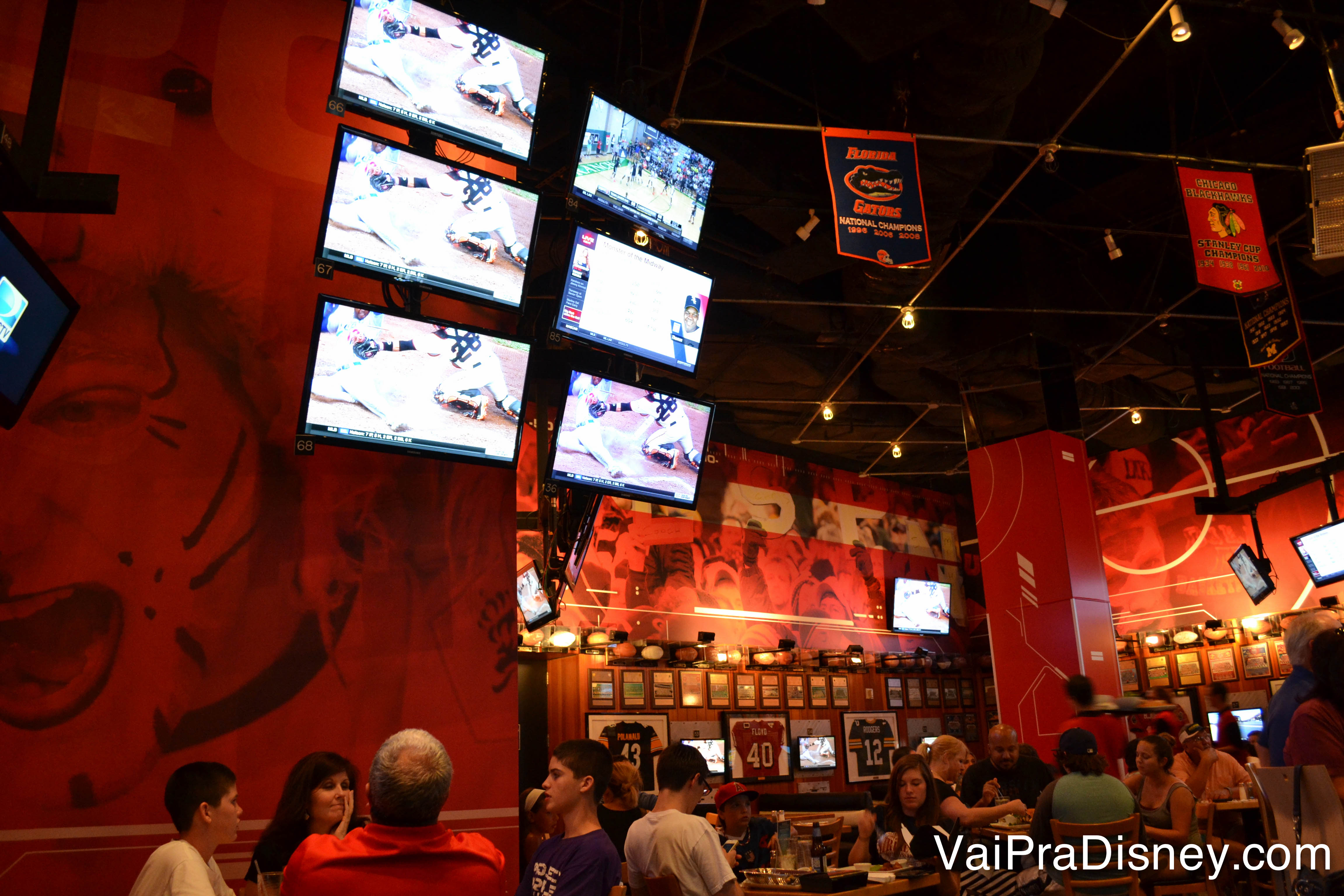 Foto do interior do restaurantes, com muitas TVs ligadas passando esportes perto das mesas 