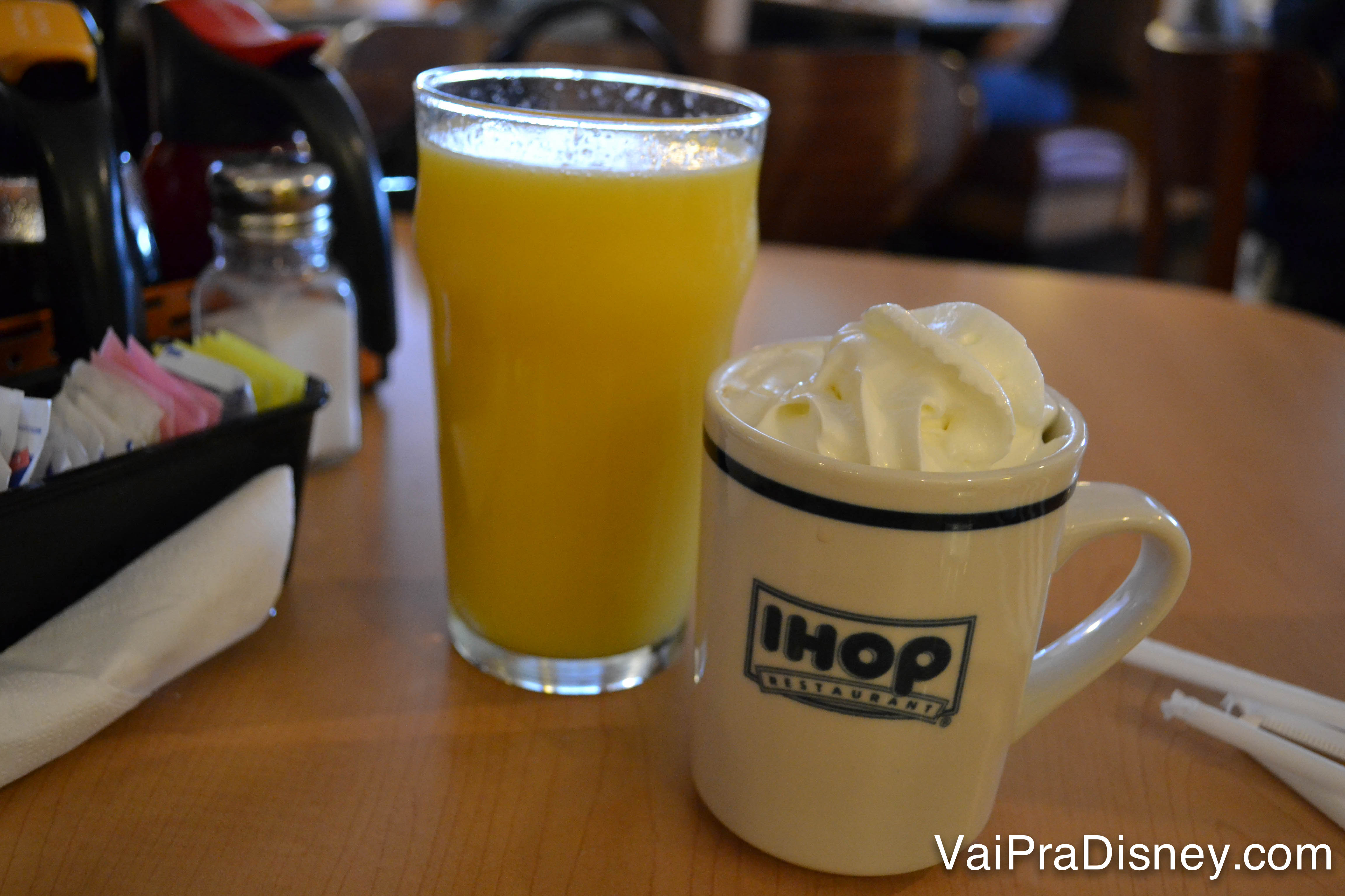 Foto de um suco de laranja no copo, ao lado de uma xícara de chocolate quente com chantilly em cima 