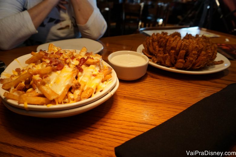 Qual você gosta mais? Foto de uma mesa com um prato de batatas fritas com queijo e bacon e um prato de Onion Rings 