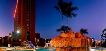 Foto da piscina do Wyndham Garden Lake Buena Vista, que tem pedras e palmeiras ao seu redor. Ao fundo é possível ver o prédio do hotel e o céu escurecendo.