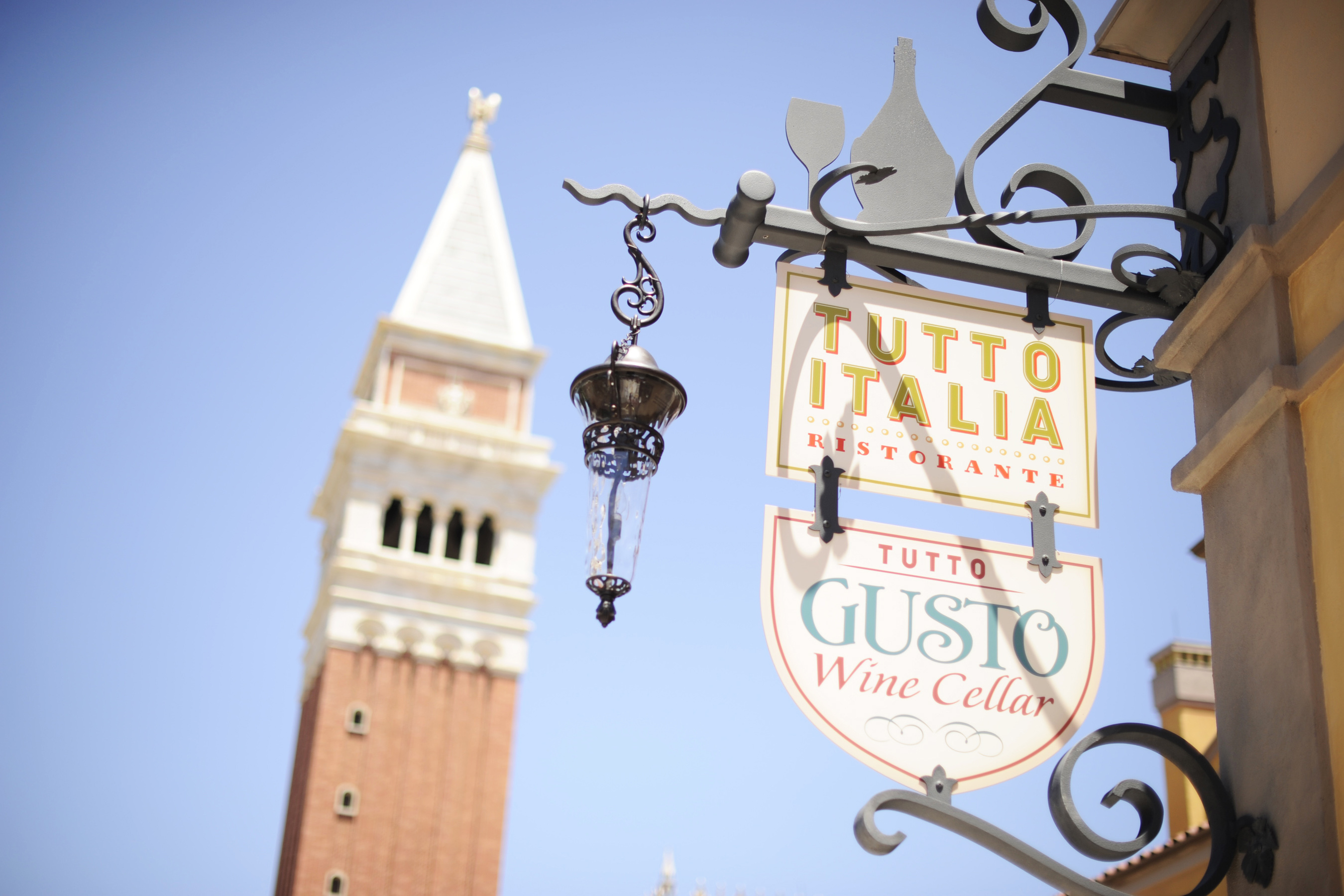 Foto da placa do Tutto Italia, com a torre do pavilhão da Itália no Epcot ao fundo e o céu azul 