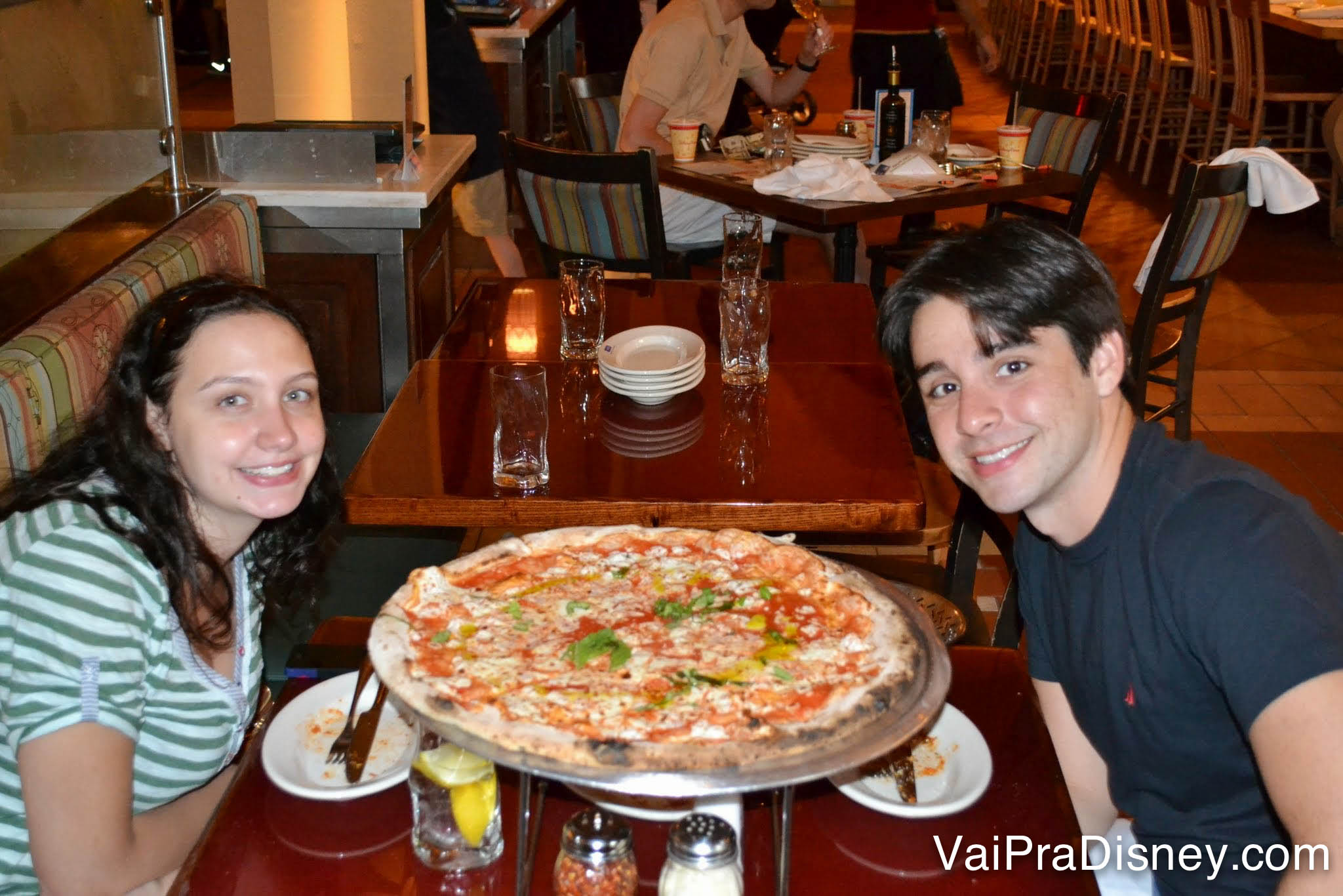 Renata e Felipe sentados à mesa com a pizza do Via Napoli entre eles 