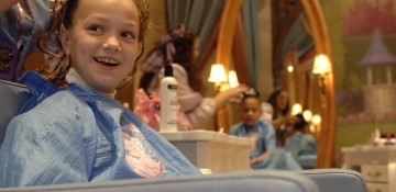 Foto de uma menina durante a transformação em princesa no Bibbidi Bobbidi Boutique. Há alguém arrumando seus cabelos, ela está com uma roupa azul e há um espelho oval no fundo da foto.