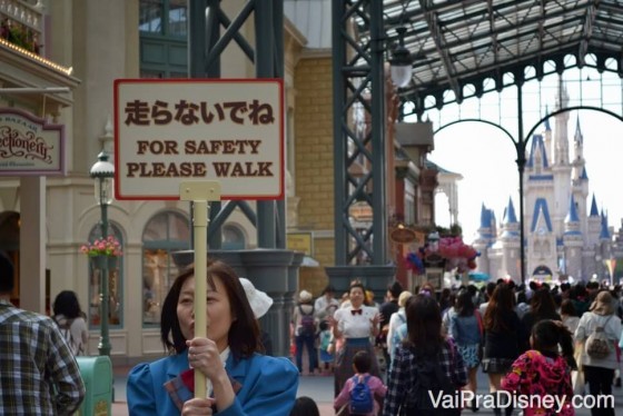 Foto de uma funcionária segurando uma placa em japonês e inglês ("for safety please walk") na Disney de Tóquio, com o castelo ao fundo e visitantes passando 