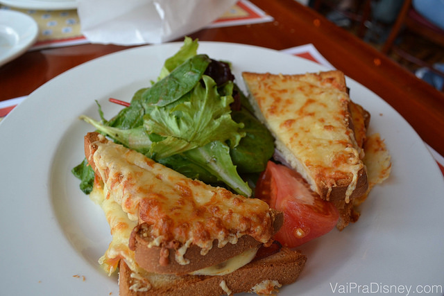 Imagem de um Croque Monsieur no prato, com queijo tostado cobrindo o pão e alface e tomate entre as duas fatias. 