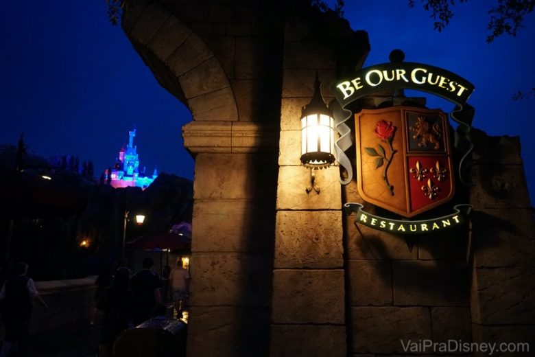 O castelo fica ainda mais bonito a noite. Foto da placa na entrada do restaurante no Magic Kingdom, durante a noite, com o castelo da Cinderela no fundo da foto. A placa iimita um brasão medieval com uma rosa, um leão e espadas.