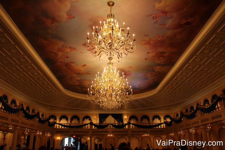 A Disney caprichou nos detalhes desse castelo! Olha que lindo! Foto da decoração do interior do castelo da Fera, com um lustre elaborado e pinturas no teto. 