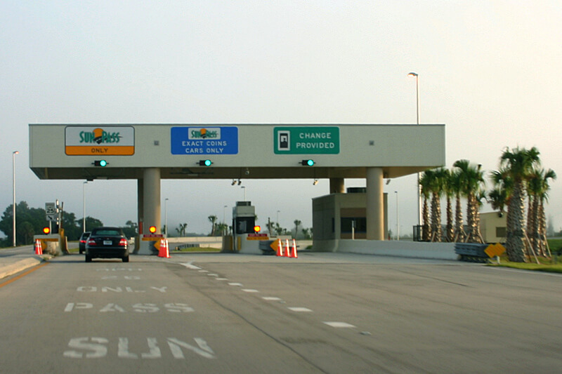 Veja os 3 tipos de cobrança em pedágios de Orlando. Assim você consegue dirigir pelos EUA. Foto de uma estação de cobrança de pedágio na rodovia em Orlando,, com diversas placas e um carro passando 