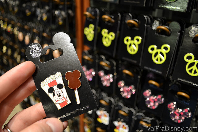 Foto dos pins menorzinhos que são vendidos em quase todas as lojas da Disney e existem milhares de modelos disponíveis, inclusive trazendo temas como aniversário e outras comemorações. Na foto, uma mão segurando um pin de pipoca e sorvete do Mickey. 