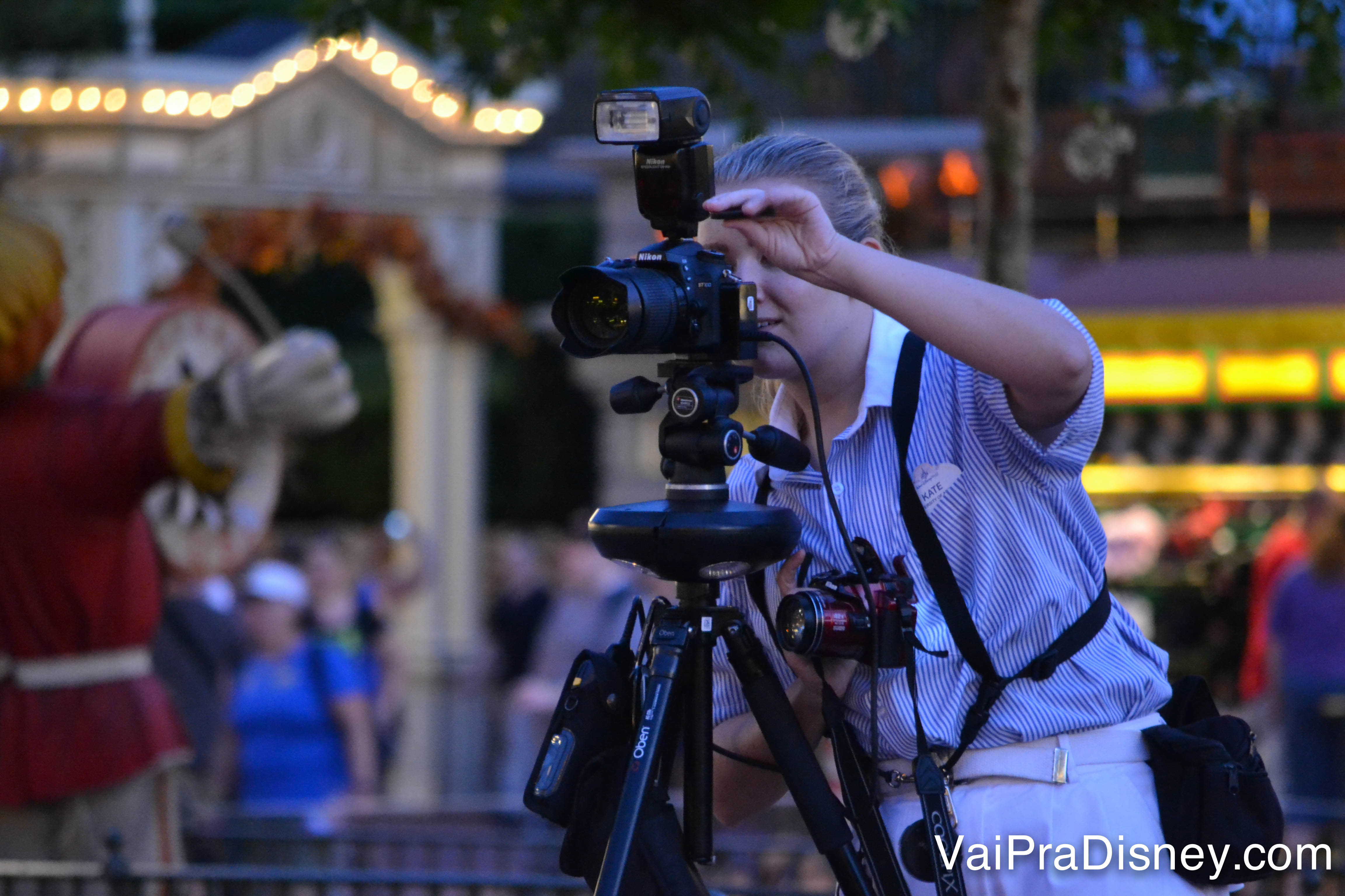 Esses são os fotógrafos do Photopass, é fácil de localizar com as câmeras e uniforme. Foto de um dos fotógrafos do Disney Photopass, atrás da câmera no parque enquanto fotografa um visitante. 