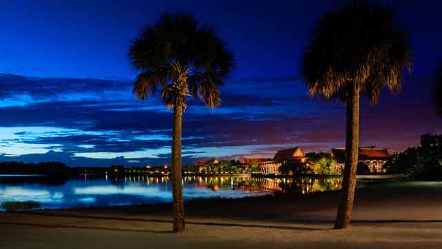 Foto de divulgação da Disney do Disney's Polynesian Resort, visto de longe, todo iluminado com a praia e as palmeiras ao seu redor. 