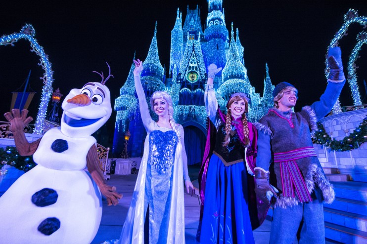 Personagens de Frozen iluminando o castelo no Frozen Holiday Wish. Foto da rainha Elsa, princesa Anna, Olaf e Kristoff em frente ao castelo da Cinderela no Magic Kingdom, iluminado em tons de azul contra o céu escuro.