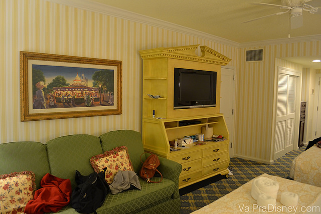 Não repara a bagunça no nosso sofá, tá? ;) Foto do quarto do hotel, mostrando o móvel de TV amarelo e um sofá verde com mochilas em cima. 