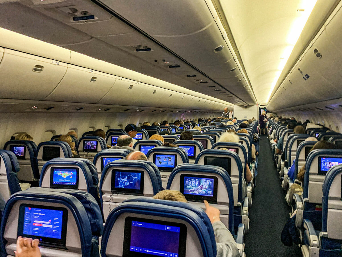 Imagem do interior de um avião. É possível ver a parte de trás de diversas fileiras de poltronas, com as telas acopladas atrás.