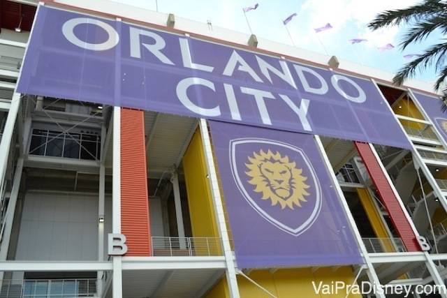 Foto do lado de fora do estádio, com as cores e o brasão do Orlando City (um leão amarelo no fundo roxo) 