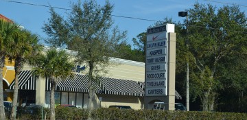 Foto da entrada do Lake Buena Vista Factory Stores, com uma placa grande com o nome das lojas presentes, algumas árvores e o céu azul ao fundo