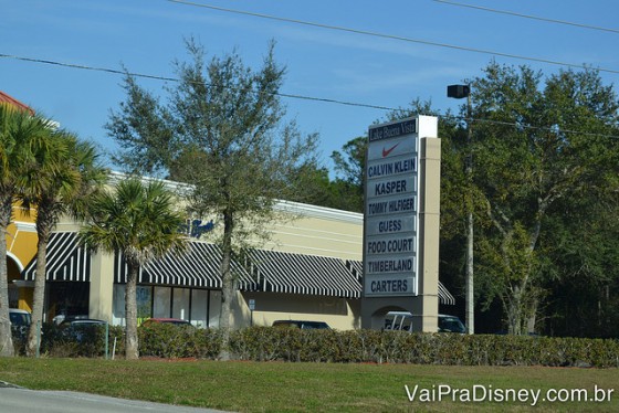 Foto da entrada do Lake Buena Vista Factory Stores, com uma placa grande com o nome das lojas presentes, algumas árvores e o céu azul ao fundo