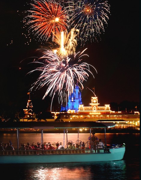 Foto dos fogos do Magic Kingdom, com o castelo e o parque iluminados ao fundo, e o  barco do Pirates & Pals Fireworks Voyage em primeiro plano 