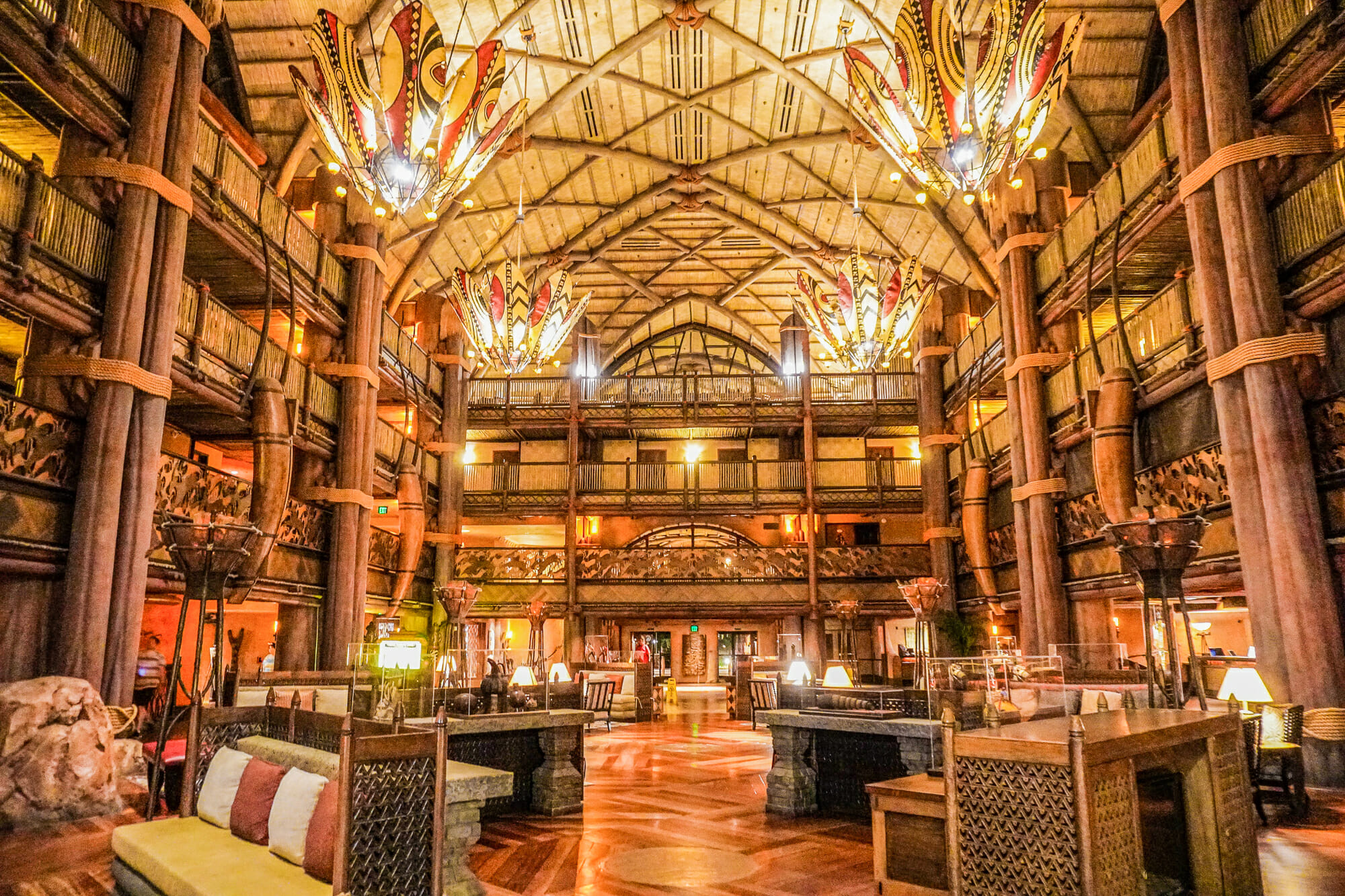 A imagem mostra o lobby do Animal Kingdom Lodge, um dos hotéis de luxo da Disney. A decoração é toda em madeira, com pilares, teto alto e lustres coloridos.