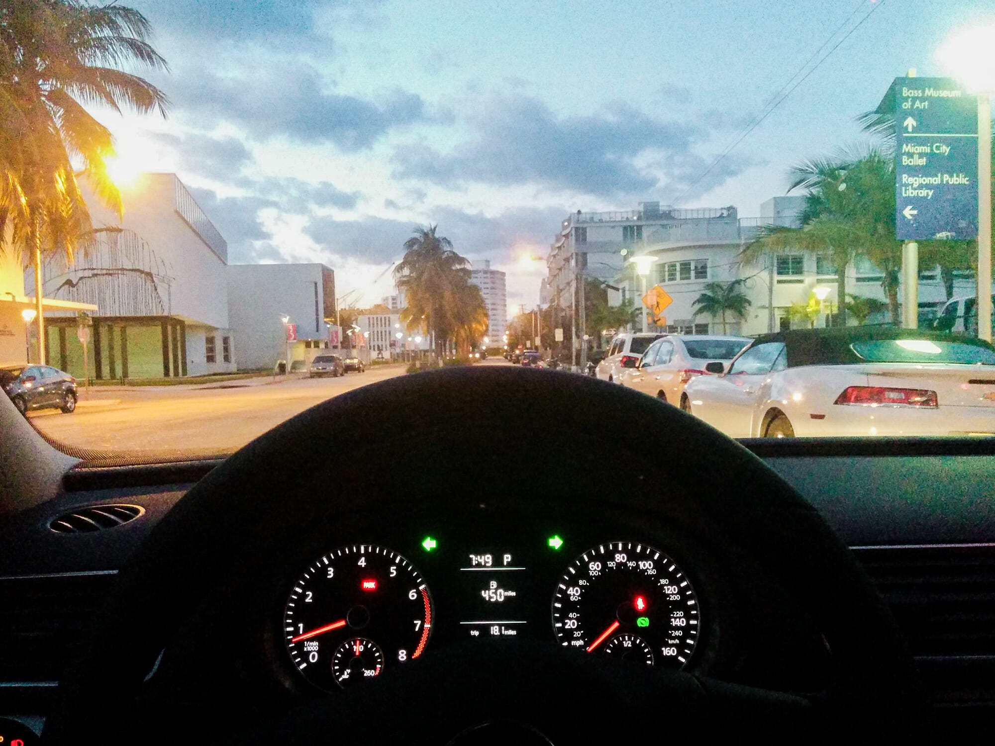 Foto tirada de trás do volante do carro, mostrando uma cidade ao entardecer, com algumas luzes já acesas