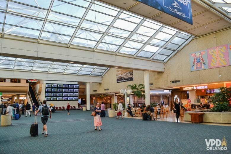 Foto do saguão do aeroporto de Orlando. É possível ver algumas lojas, passageiros e uma escada rolante. 