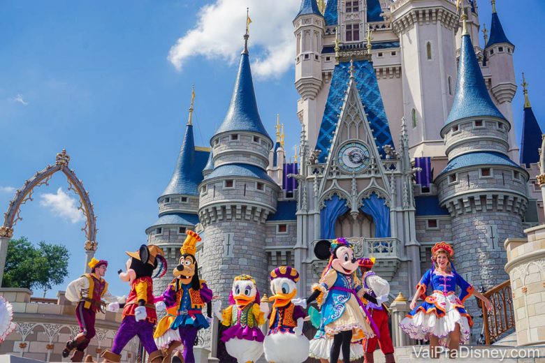 Foto de diversos personagens como Minnie, Mickey, Donald e Pluto em frente ao castelo da Cinderela no Magic Kingdom, com o céu azul ao fundo. 