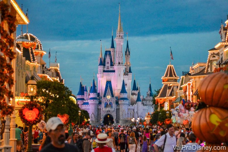 Foto da Main Street do Magic Kingdom cheia de visitantes, decorada para o Halloween, com as luzes começando a se acender, o céu escurecendo e o castelo da Cinderela ao fundo. 