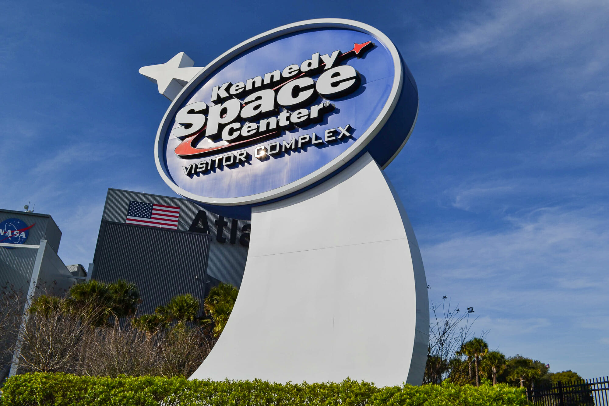 Foto da placa na entrada do Kennedy Space Center, que tem o logo do local e um foguete branco ao lado.