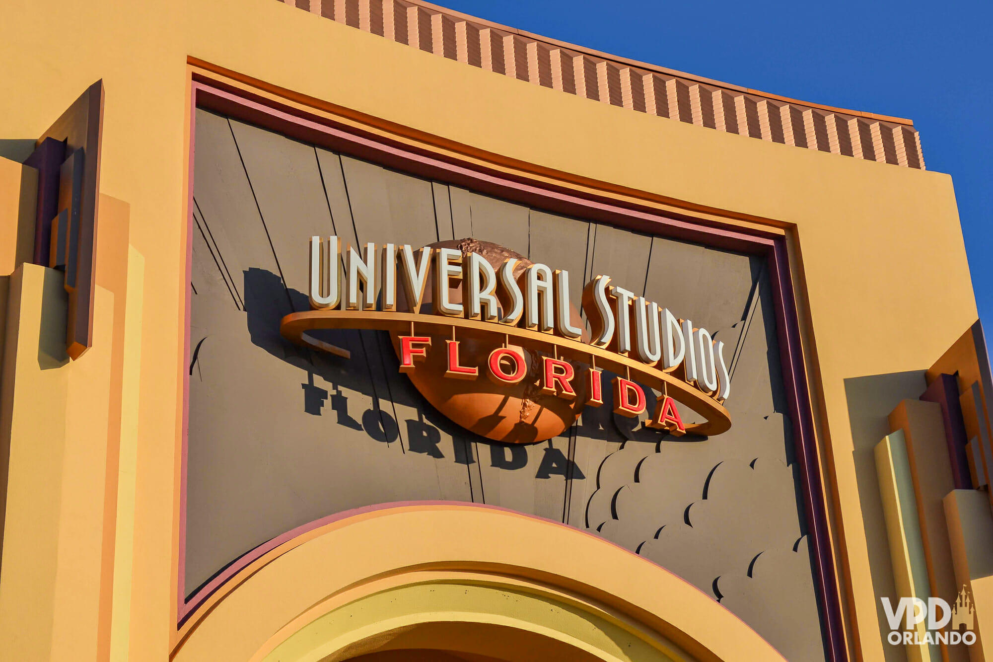 Foto do portal do parque Universal Studios Florida, que mostra a placa com o nome do parque escrito em branco e laranja. O portal é pintado em bege e laranja-claro e o céu ao fundo está bem azul.