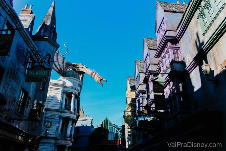 Tem MUITA atração legal do Harry Potter na Universal! Foto do Beco Diagonal do Harry Potter na Universal, com construções coloridas, o dragão de Gringotts e o céu azul ao fundo. 