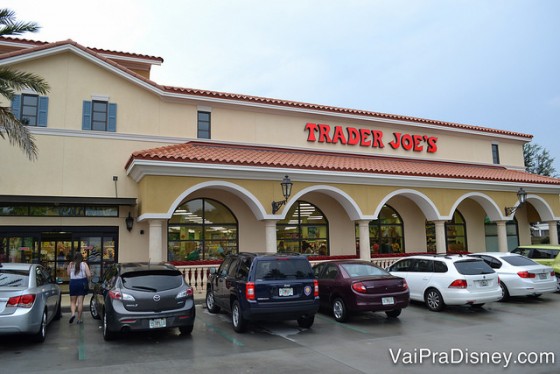 Foto do exterior do supermercado Trader Joe's em Orlando, pintado de bege claro, com o nome do local em letras vermelhas e diversos carros estacionados na frente. 