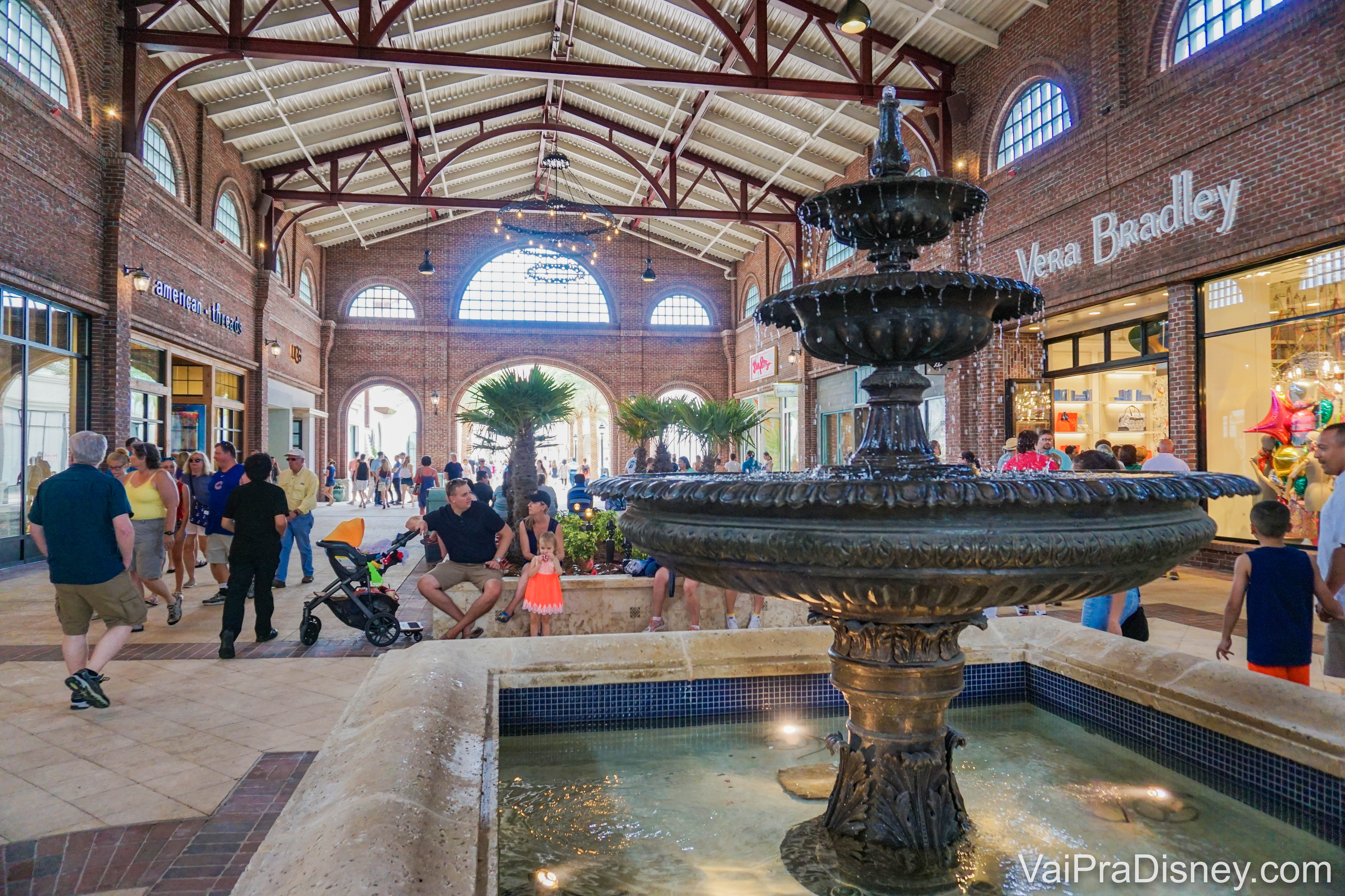 Foto das lojas em Disney Springs, em uma área coberta, com a fonte em primeiro plano. 