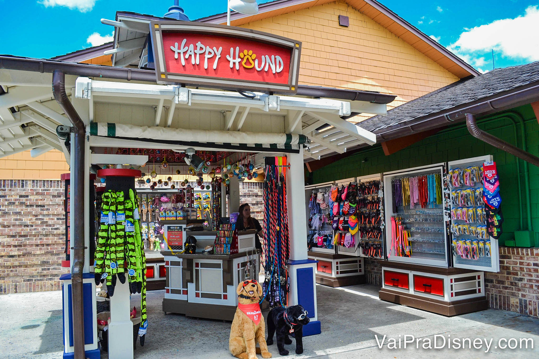 Foto do quiosque Happy Hound, que vende produtos para cachorros. Há um cachorro de pelúcia em frente, com bandana e óculos escuros. 