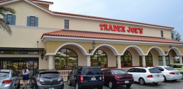 Foto do exterior do supermercado Trader Joe's em Orlando, pintado de bege claro, com o nome do local em letras vermelhas e diversos carros estacionados na frente.