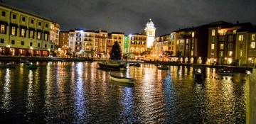 Foto do Portofino Bay iluminado durante a noite, com o lago, um barco passando e as lojas e restaurantes mais ao fundo