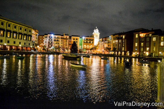 Foto do Portofino Bay iluminado durante a noite, com o lago, um barco passando e as lojas e restaurantes mais ao fundo 