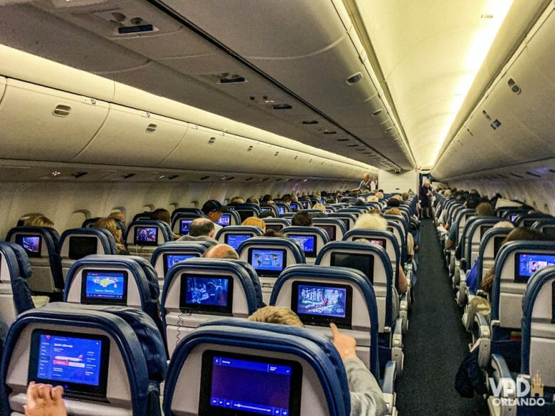 Foto do corredor no interior de um avião. É possível ver a parte de trás de diversas fileiras de poltronas, com as telas acopladas atrás. 