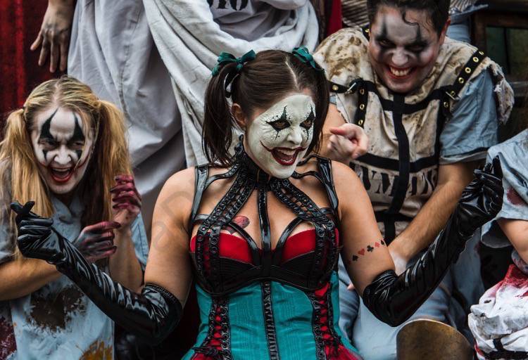 Personagens aterrorizantes na Halloween Horror Nights da Universal. A imagem mostra uma moça vestida e maquiada como uma arlequina assustadora, com personagens semelhantes atrás. 