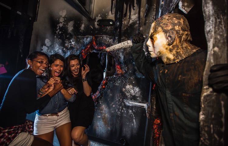 Halloween Horror Nights da Universal. A imagem mostra três visitantes levando um susto com uma pessoa vestida de Michael Myers, personagem de Halloween, com uma faca na mão. 