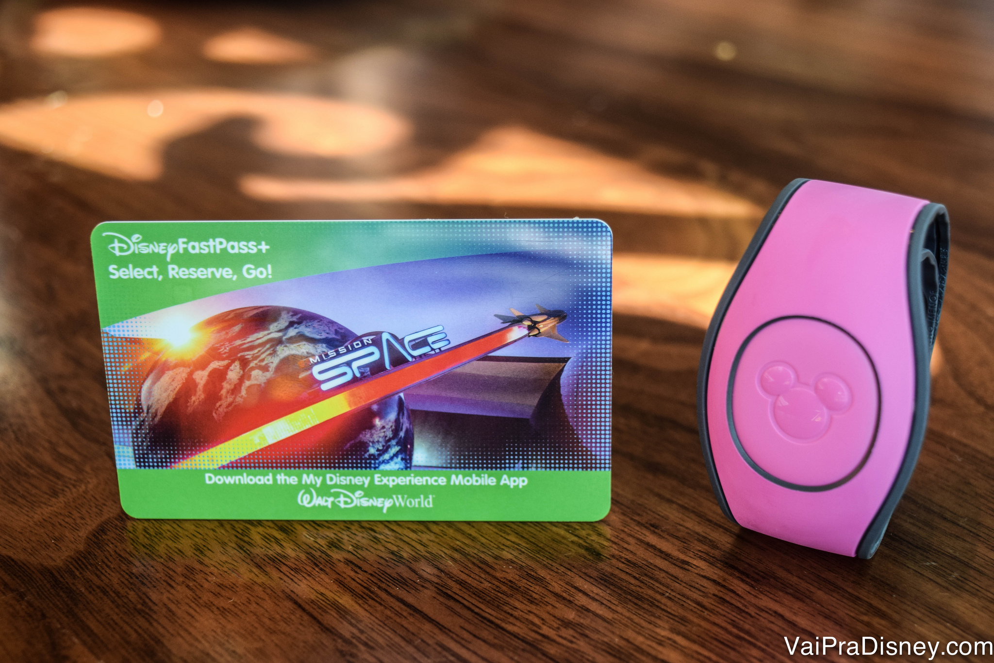 O ingresso pode ser usando em formato de cartão ou com a MagicBand. Imagem do cartão magnético que funciona como ingresso da Disney, ao lado de uma MagicBand cor-de-rosa.