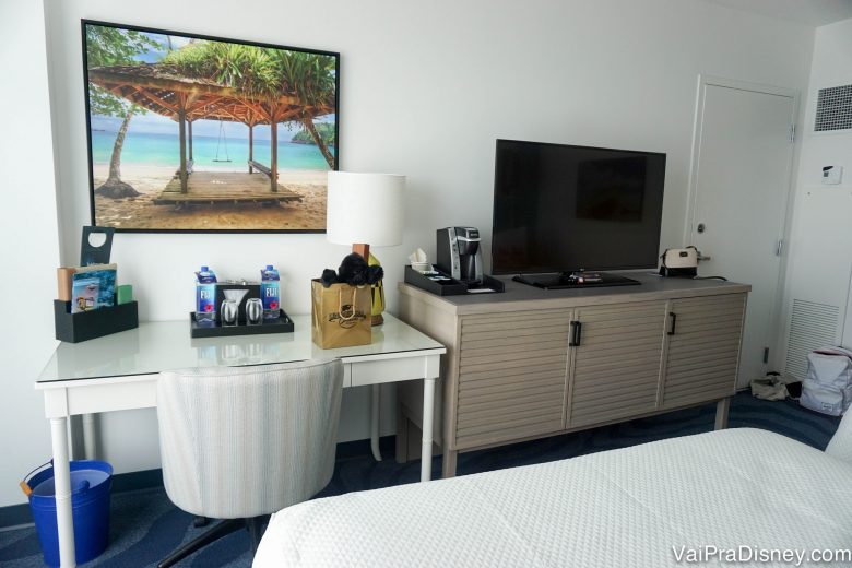 Foto do quarto mostrando um quadro de praia, uma escrivaninha e um móvel com tv em cima 