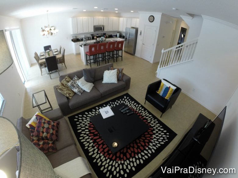 Outro ângulo da casa para poder dar noção do espaço. Foto de uma sala de estar com cozinha e sala de jantar ao fundo. A sala tem sofás cinzas, tapete e mesa de centro. 