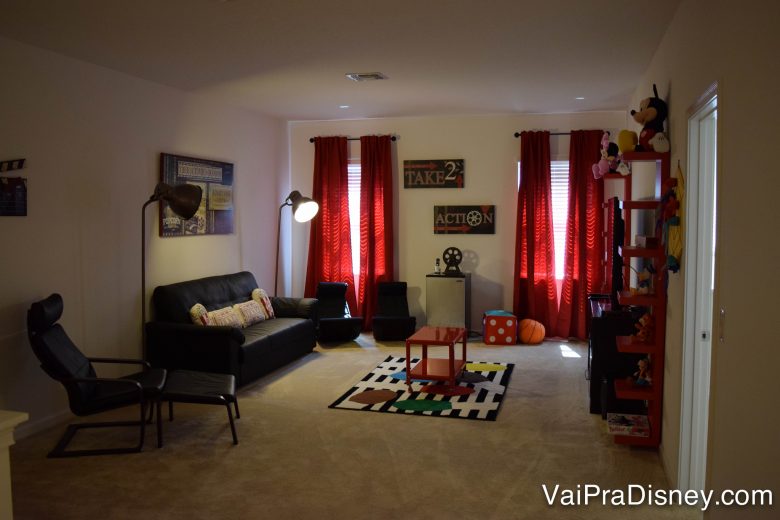 Salinha de TV no segundo andar da casa. Foto da sala de TV na casa alugada, com um sofá preto, uma poltrona preta e cortinas vermelhas. 