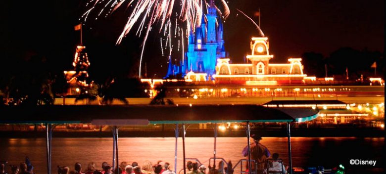 Foto do Magic Kingdom, com o castelo iluminado ao fundo e parte do lago e de um barco com visitantes visível em primeiro plano.
