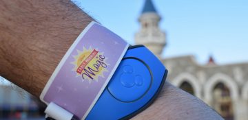 Foto de uma mão com uma MagicBand azul no pulso e uma pulseira lilás identificando a pessoa como participante do Early Morning Magic. É possível ver o castelo da Cinderela ao fundo da foto.