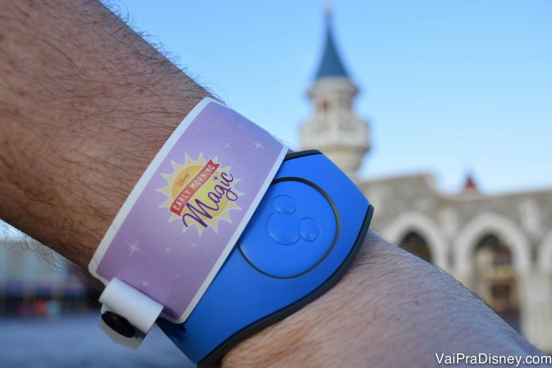 Foto de uma mão com uma MagicBand azul no pulso e uma pulseira lilás identificando a pessoa como participante do Early Morning Magic. É possível ver o castelo da Cinderela ao fundo da foto.