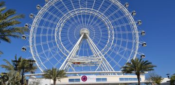 Foto da roda-gigante conhecida como The Wheel, em Orlando, com o céu azul ao fundo e a entrada do ICON Park em primeiro plano.