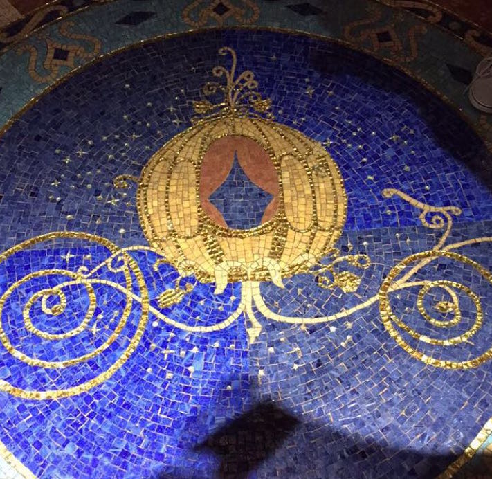 O chão do hall é um dos detalhes mais lindos da suíte, junto com o sapatinho que também fica por lá. Foto do mosaico no chão do hall, que mostra a carruagem da Cinderela em dourado sobre um fundo azul. 