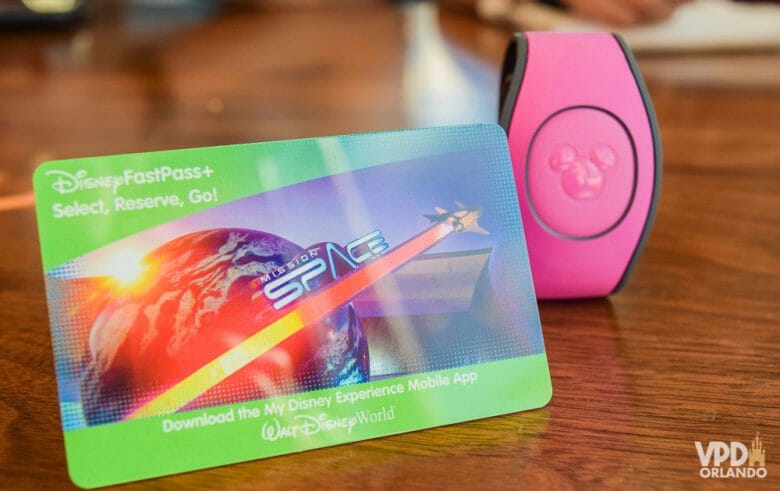 Foto do cartão magnético que serve como ingresso dos parques da Disney, ao lado de uma MagicBand cor-de-rosa. 