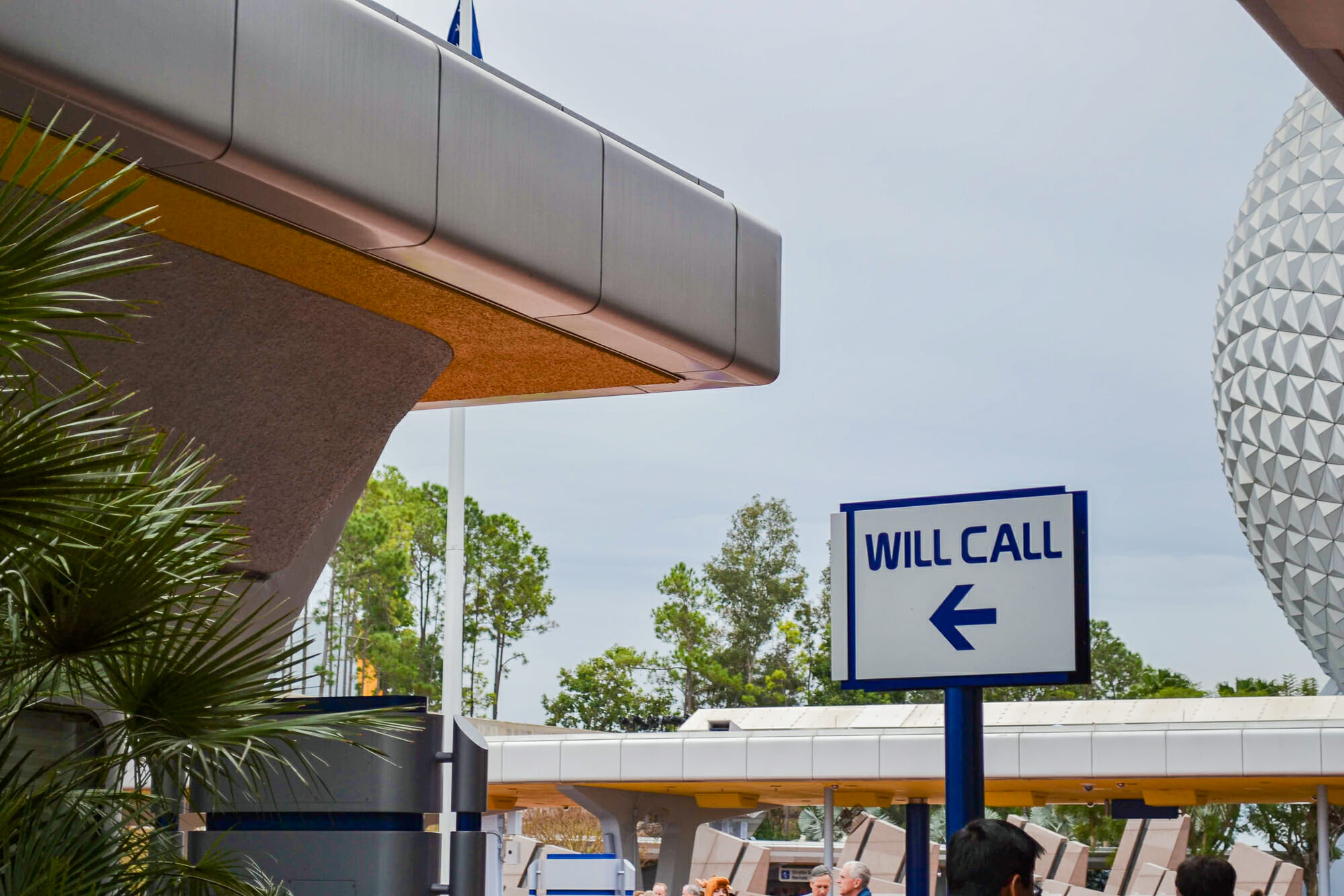 Foto com a placa indicando a direção do Will Call no Epcot, com parte da "bola" visível na lateral da imagem. A placa tem fundo branco, letras azuis, uma flecha apontando para a esquerda e diz "Will Call"